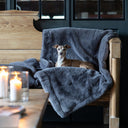 A dog lying on a plush faux-fur dog blanket in a grey blue colour called Russian Blue draped on a wooden bench chair in a home setting.