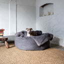 A dog lying in a cosy donut calming dog bed with a grey blue luxury faux-fur dog blanket in a home setting with whitewashed walls and a hessian floor.