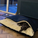 Whippet dog lying in a cave bed style Snuggle Bed in navy velvet with a cream lining  in a room with wooden flooring and a sliding glass door.