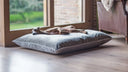 A cool and comfortable dog lying stretched on a large grey luxury dog bed mattress in grey velvet in a home setting with large windows.