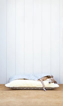 A dog sleeping inside a cool cotton sleeping bag style dog bed, The Charley Chau Snuggle Bed, with a grey polka dot outer cover and creamfluffy lining with a panelled wall behind and natural flooring.