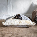A black dog sleeping cosy and covered up inside a luxury Dog Snuggle Bed with a cream faux-fur lining in a home setting with a whitewashed wall and hessian natural carpet.