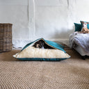 A black dog sleeping under cover in  a cave bed style dog bed in green with a cream faux-fur lining in a home setting with a whitewashed wall and hessian natural carpet.