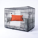 A wire dog dog crate with a luxury crate mattress and bed bumper set in pale beige/grey with a small orange  mini-bolster pad cushion on a white backdrop.