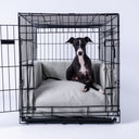 A black Italian Greyhound lying inside a dog crate very comfortable on a pale grey/beige luxury dog crate mattress with matching crate bed bumpers on a white backdrop.
