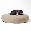 A black dog curled up very comfortable and calm on a beige colour round luxury dog bed mattress on a white backdrop. 