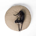 A black dog lying on a beige coloured round luxury dog bed mattress seen from above on a white backdrop. 
