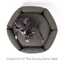 A black dog lying in a luxury donut dog bed see from overhead in a woven upholstery fabric in dark brown with high bed sides and a deep-filled luxury mattress on a white backdrop.
