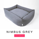 A cool blue-grey Bliss Bolster Dog Bed in 'NIMBUS GREY' Faroe fabric shown from an angle on a white background, highlighting the tall sides of this long lasting dog bed with sides; a stylish bolster dog bed and high-wall dog bed for dogs of all sizes.