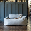 A dog lying in a box bed style bolster bed with high sides in a pale grey in a stylish home setting with wood panelled wall and dark wooden floor.