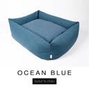 A deep petrol blue Bliss Bolster Dog Bed in 'OCEAN BLUE' Faroe fabric shown from an angle on a white background, highlighting the tall sides of this long lasting dog bed with sides; a stylish bolster dog bed and high-wall dog bed for dogs of all sizes.