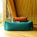 A dog lying in a jade green luxury high-sided Bolster Dog Bed in a hard-wearing outer fabric, with two small cushions inside the bed that the dog is leaning against, in a home setting with wood panelled walls and a wooden floor.
