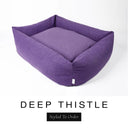 A rich deep purple Bliss Bolster Dog Bed in 'DEEP THISTLE' Faroe fabric shown from an angle on a white background, highlighting the tall sides of this long lasting dog bed with sides; a stylish bolster dog bed and high-wall dog bed for dogs of all sizes.