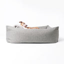 A dog lying in a box style luxury dog bed with high sides by designers Charley Chau on a white backdrop. 