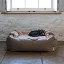 A dog sleeping very cosy with a beige sherpa fleece dog blanket in a high-sided bolster dog bed in abeige made with a durable upholstery fabric in a home setting with a whitewashed wall and stone floor.