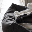 Close up of a dog sleeping very content in a luxurious bolster dog bed made with a durable upholstery fabric,  with a pale cream faux-fur dog blanket in a home setting.