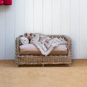 Luxury Raised Rattan Dog Bed on a hessian carpet with a aple grey panelled wood wall. with a beige deep-filled dog bed mattress and a dog sleeping under a stylish faux-fur dog blanket.